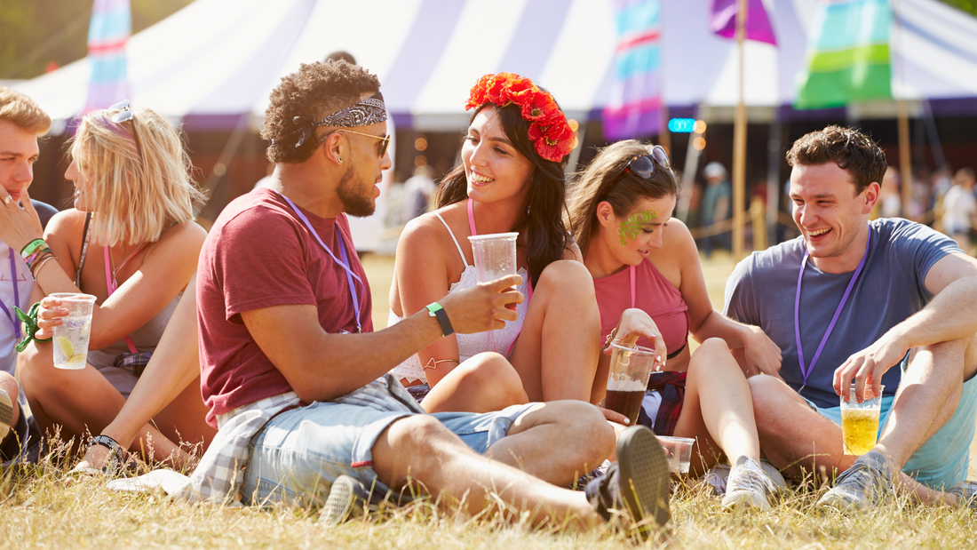 A group of people sitting on the ground having drink at a festival. One lady is wearing a headband covered in poppies and flowers