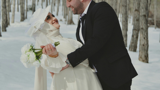 A groom looking at his bride in a snow covered forest. She's wearing a hat.