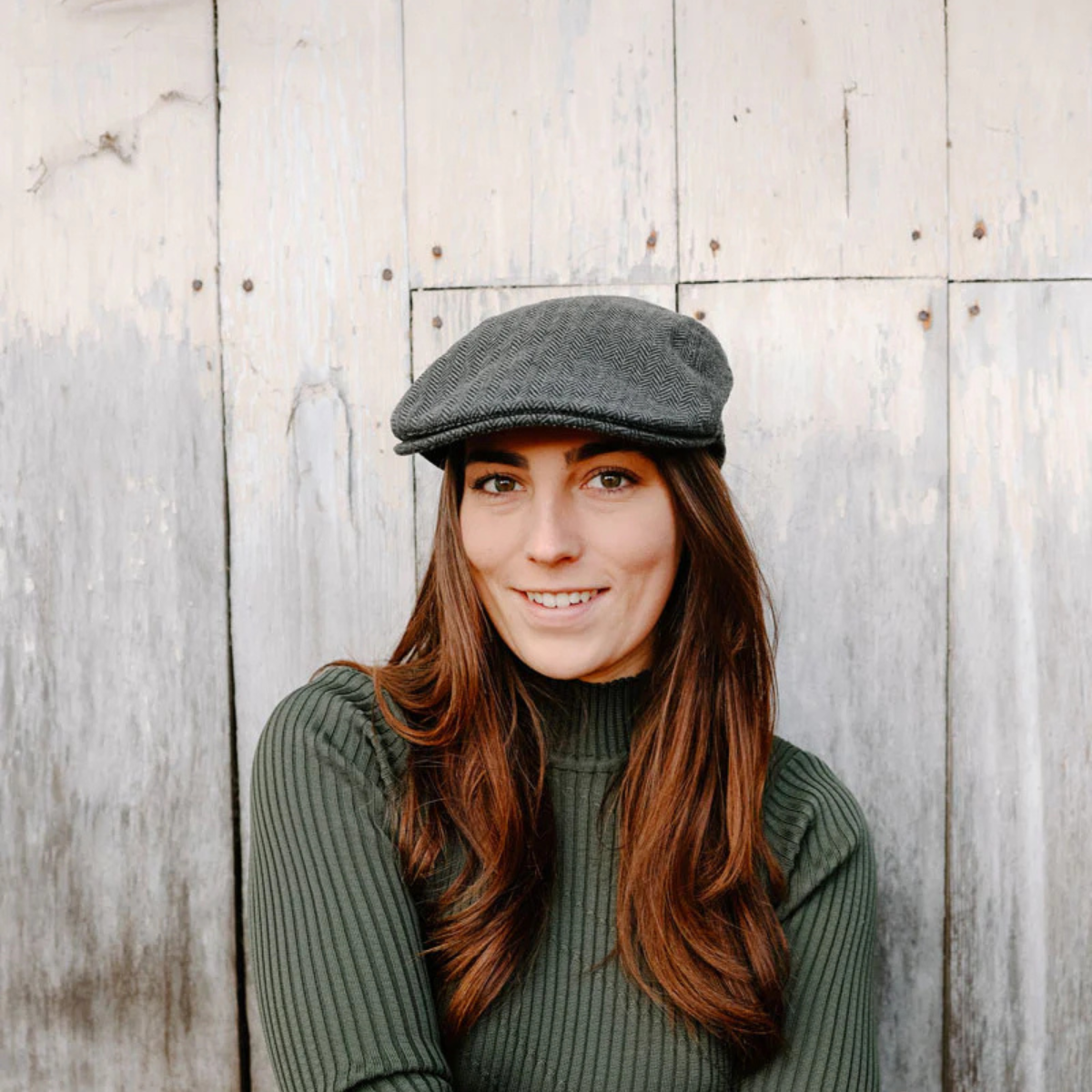 A lady standing in front of a wooden building wearing a flat cap